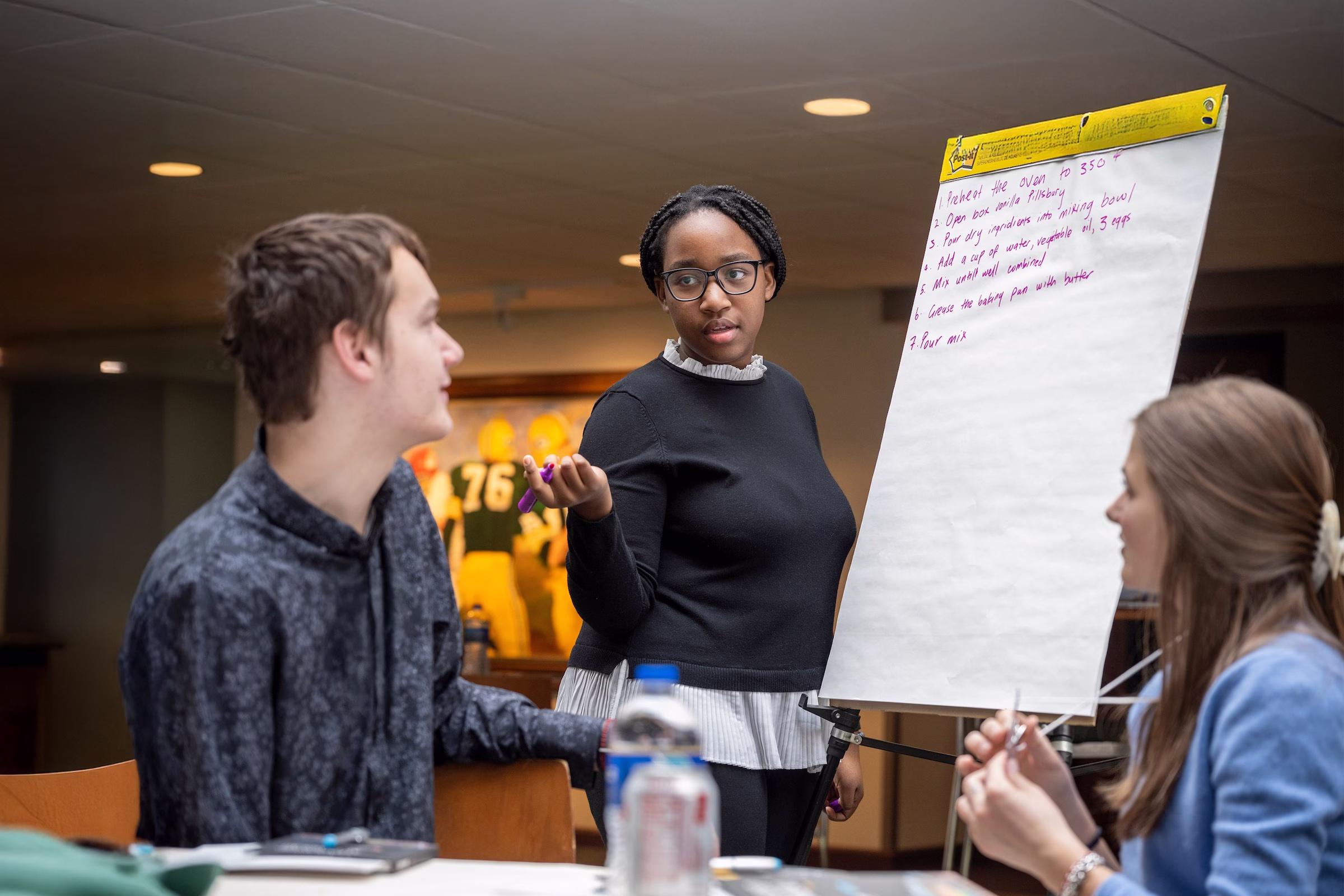 A woman with a large poster paper talking with people sitting at a table.