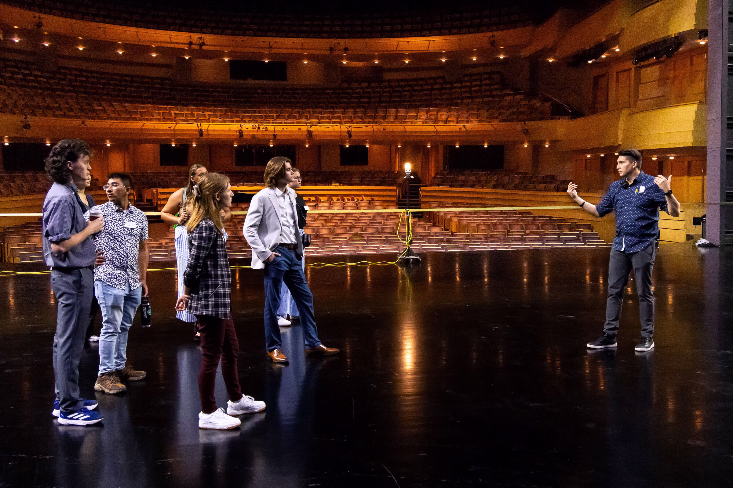 Students standing on a stage with one person talking in front of them.
