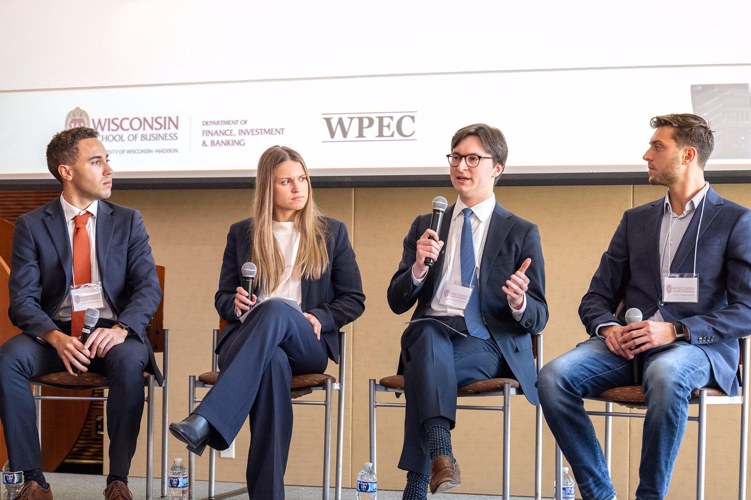 Four students sit on stools on a stage during a panel discussion; one student is speaking into a microphone.