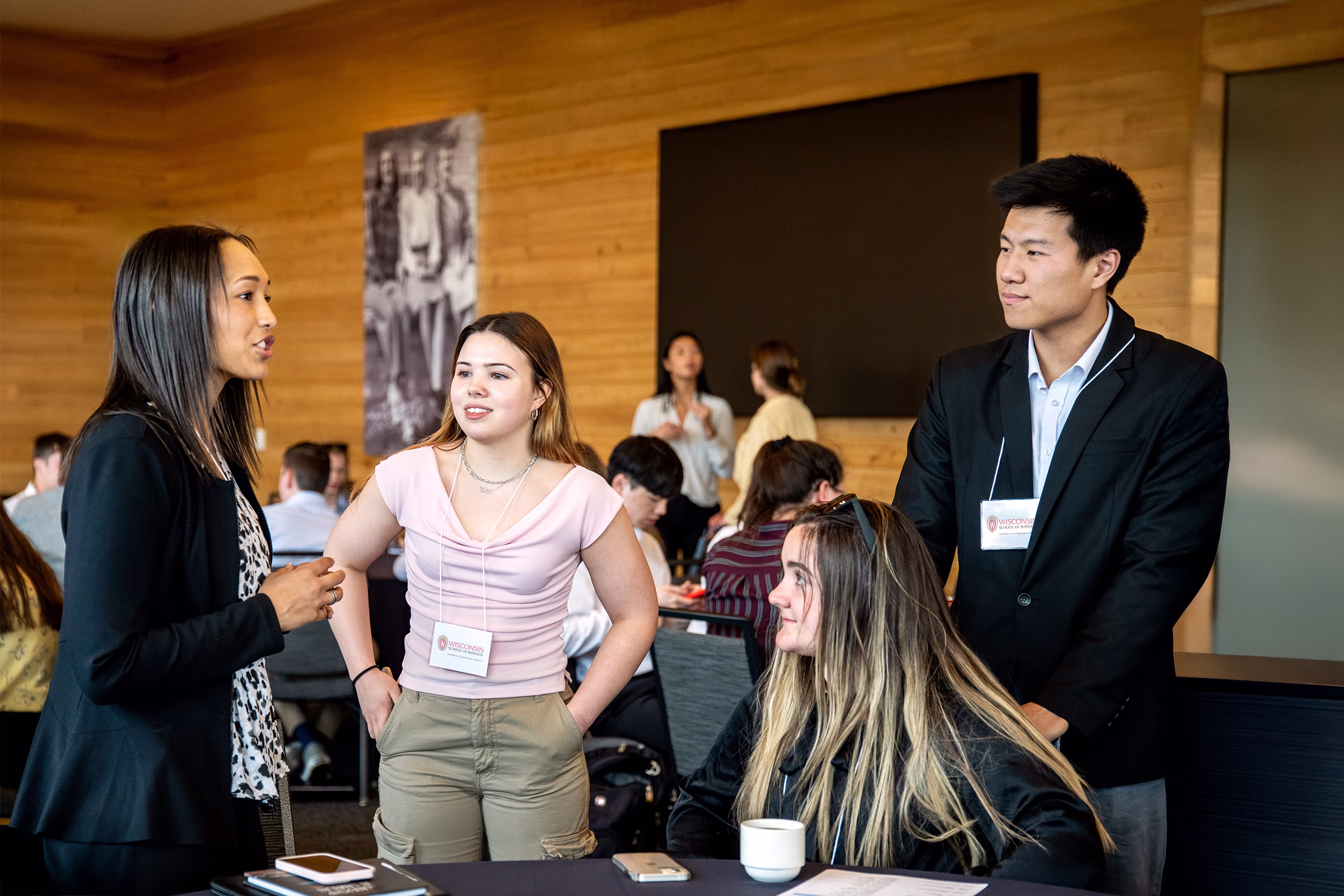 Three students stand in a group with another student sitting at a table.