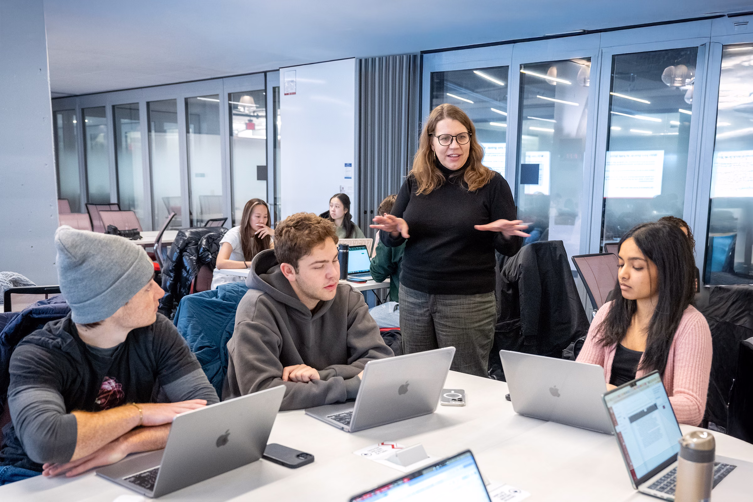 Senior lecturer Katie Krueger stands and chats with three students sitting at a table with their laptops open in front of them.