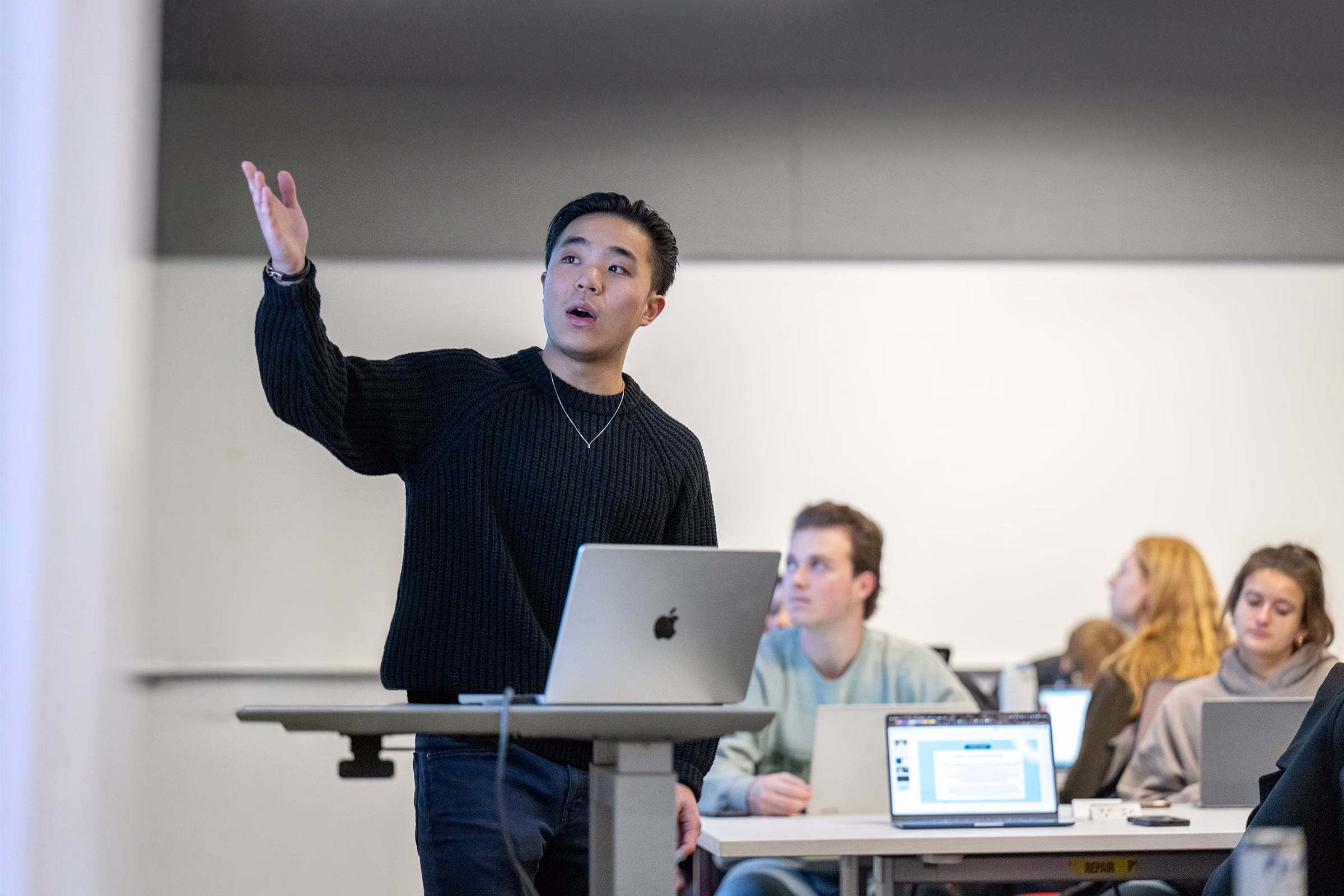 A male student points to a slideshow while he stands and presents to a class of his peers.