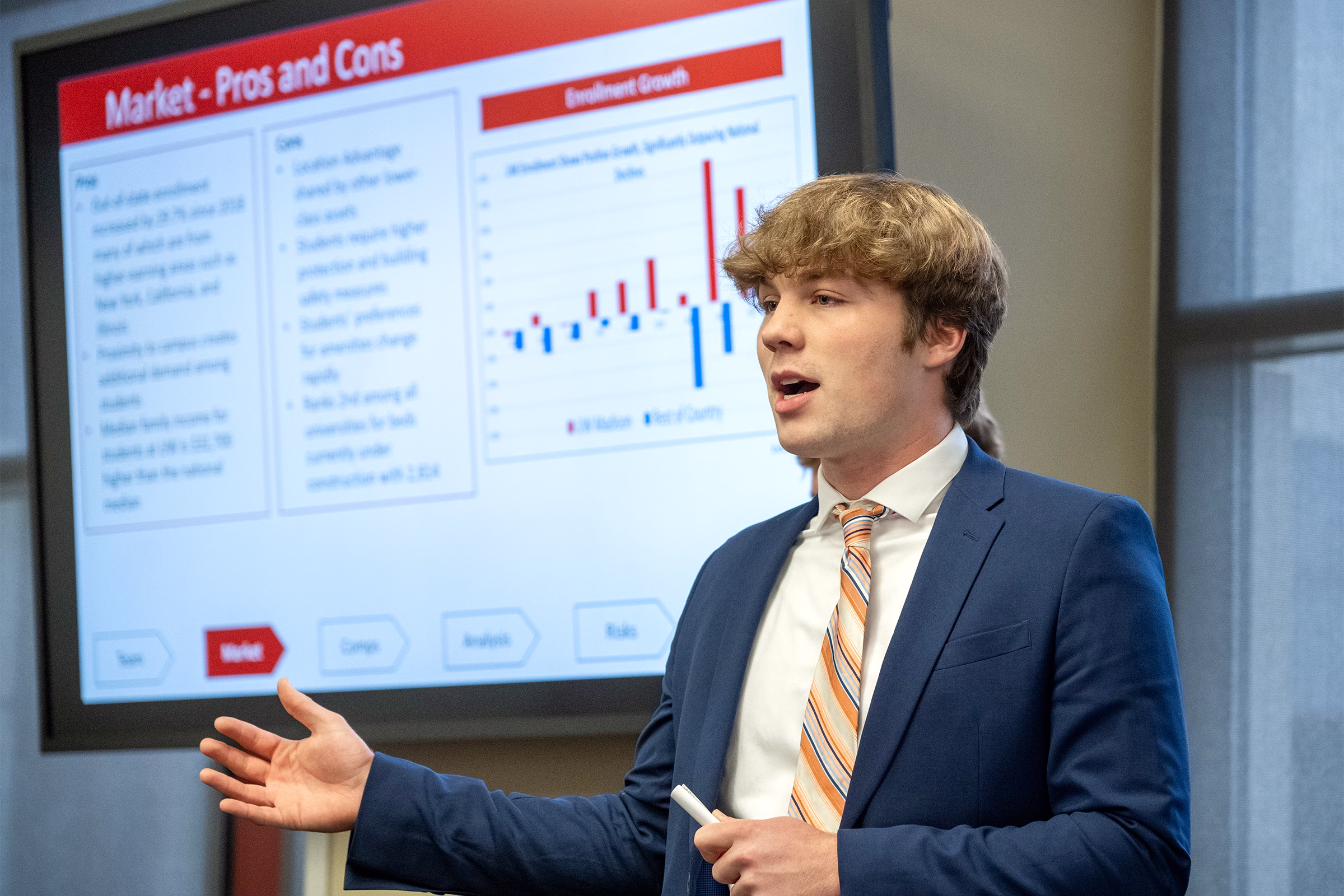 A male student wears a suit and tie while presenting in front of a slideshow at a case competition.