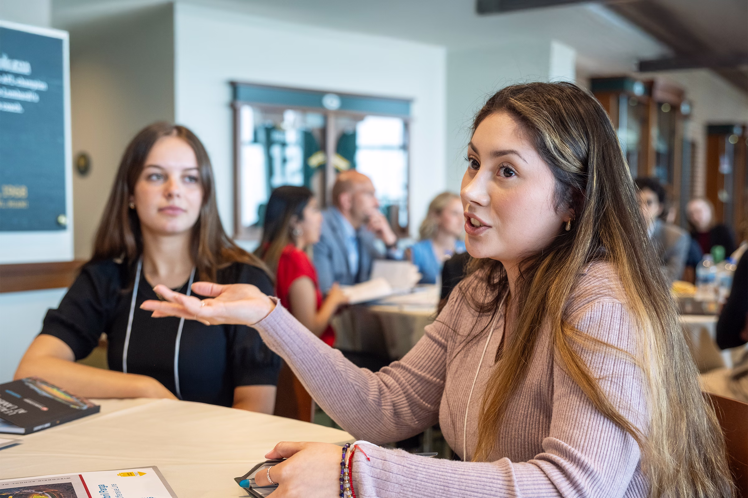 Two female students sit at a table with a group while one of them talks.