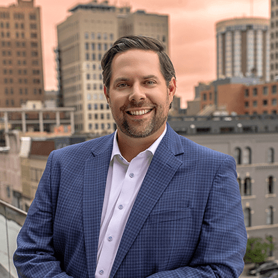 Jason Eggert in a suit, smiling in front of backdrop of tall buildings.