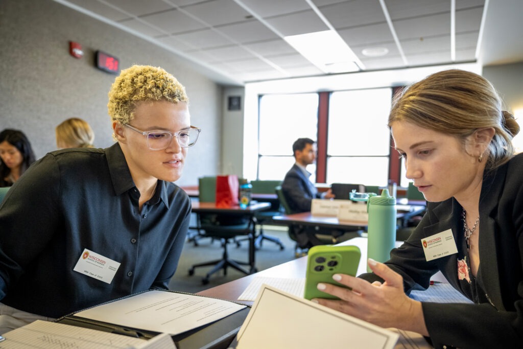 Two students sit at a table while one shows the other her phone screen.