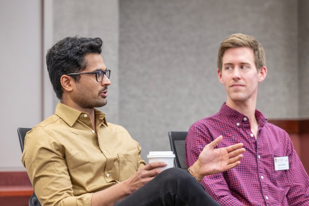 Two male students sit in a class, one of them is talking and holding a coffee cup.