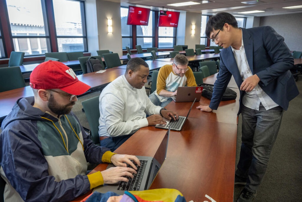 A man standing with his hand on a table and three people sitting at the table with their laptops open on the table.