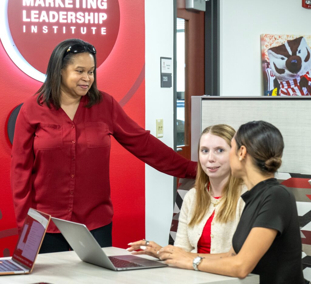 Two students sit and talk at a table while an advisor stands nearby.