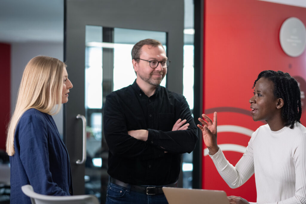 3 people standing in a group. 1 women talking and gesturing with hands.