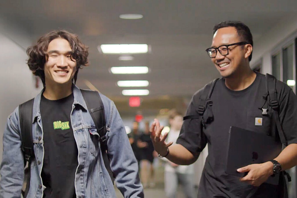 Two students smile and talk while walking down a hallway.