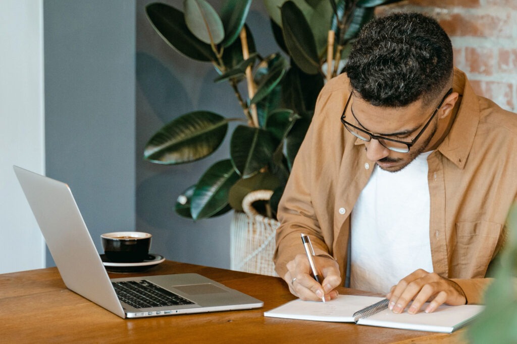 Man writing in his note book with his laptop open.