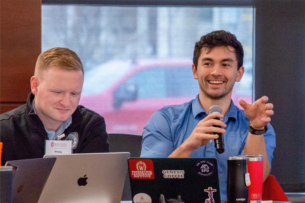 Two men sitting at a table with laptops open in front of them, and the man on the right is speaking in a microphone.
