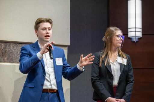 Two students stand on stage presenting, while one holds and speaks into a microphone.