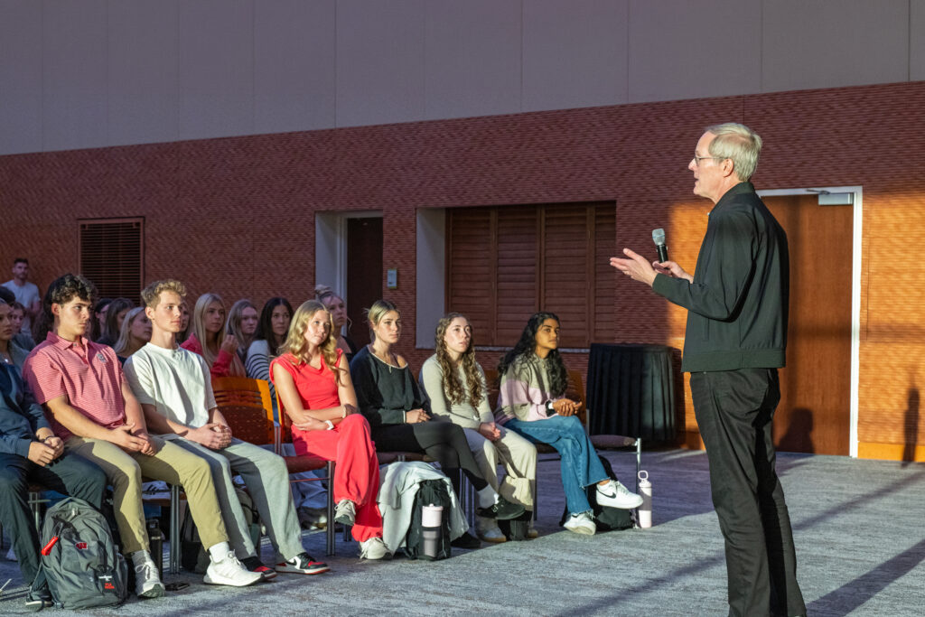 Students sit in chairs, listening to a speaker who is standing and talking into a microphone.