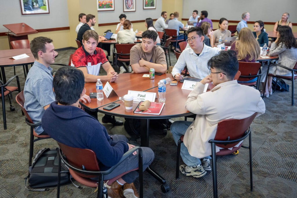 A group of students sitting at a table listening to person on the far left.