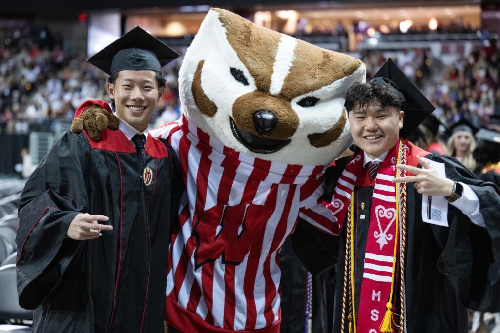 Two BBA graduates in their gowns posing with Bucky Badger.