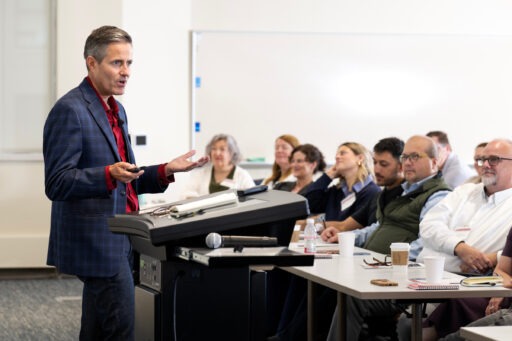 A person in a blazer stands at a podium addressing an audience seated at tables in a conference room.